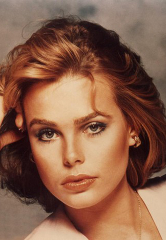 Margaux Hemingway con capelli ondulati fino alle spalle, indossa orecchini e una camicetta chiara, posa con una mano vicino alla testa su uno sfondo da studio illuminato delicatamente, esprimendo un tono calmo e introspettivo
