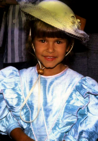 Young girl wearing a light blue satin dress with puffed sleeves and a wide-brimmed pale yellow hat adorned with ribbon and flowers, standing indoors in front of a person dressed in striped clothing. The setting appears to be a formal or celebratory event. The overall mood is gentle and nostalgic.