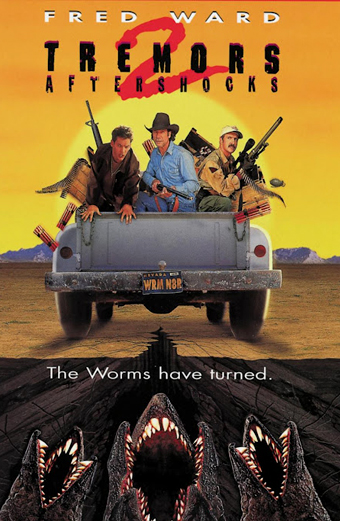 Tre uomini armati viaggiano sul retro di un pick-up che corre in un paesaggio desertico al tramonto, mentre enormi mostri simili a vermi emergono dal terreno sottostante. Il testo del poster riporta FRED WARD, TREMORS 2 AFTERSHOCKS, The Worms have turned. Il tono è teso e avventuroso, suggerisce azione e pericolo.