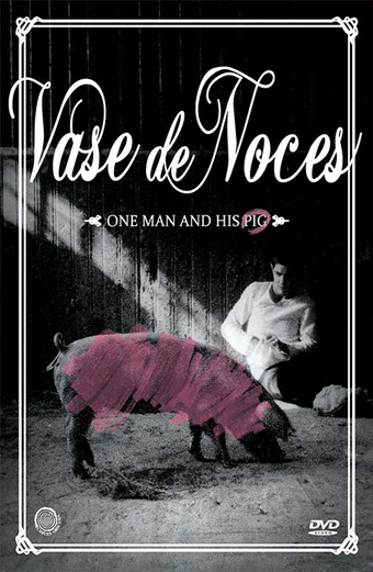 Copertina DVD di Vase de Noces. Un uomo è seduto a terra in un fienile poco illuminato, di fronte a un maiale con una striscia rosa sul corpo. L'atmosfera è cupa e surreale. Testo sul poster: Vase de Noces, One man and his pig. Una cornice bianca decora l'immagine.